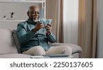 Small photo of Happy old bald overweight African American senior mature elderly man happy with financial savings salary at home sitting couch. Winning online lottery holding dollars throw up cash money