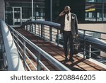 Small photo of A mature fashionable black man entrepreneur is talking on the phone while passing on an overhead passage; an African bearded businessman is phoning while standing on the pedestrian bridge with his bag