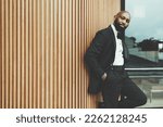 Small photo of A dandy black man with a bald head, wearing a black tuxedo, bow tie, and a white shirt; Hands in his pockets, and the other hand rests his body on a wall made of wood stakes creating an elegant design