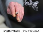 Small photo of The hand of an elderly person, holding a lit up cigarette. The nails of his index finger and ring finger are completely turned yellow because of the tar, nicotine and smoke. Concept of chain smoker.