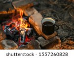 Black enamel with a cup of hot drink sits on a stone by the fire in the open air.Detail of camping.Hot coffee at the campsite, a cup at the stake and charcoal.International day of coffee
