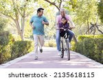 Small photo of Old man riding bicycle with granddaughter while son running at park