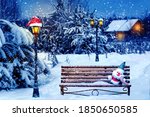 Santa hat and New Year's soft toy on a bench in the winter forest against the background of a village house and lantern. Christmas art card. Winter wonderland.