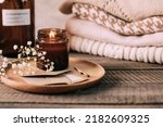 Burning candle in small amber glass jar, flowers of gypsophila and stack knitted seasin sweaters. Cozy lifestyle, hygge concept