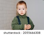 Small photo of Portrait of little cute boy in green sweater standing indoors against white wall and looking at camera. Uncomfortable facial expression, unsmiling emotion of child with big eyes, pretty baby.