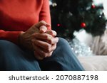 Lonely senior woman sitting at home in Christmas celebration. Close-up of an elderly woman's hand against background of decorated Christmas tree. Loneliness, sad holiday concept.