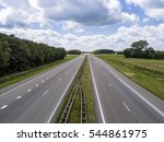 Empty motorway with no traffic except in the far distance leading straight to the horizon, in a flat green landscape with a beautiful half cloud sky