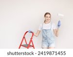 Small photo of Young attractive woman is standing at stepladder in apartment during repair works. She is holding paint roller. repair concept. House remondeling and painting.