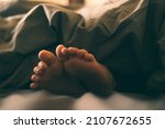 Small photo of Bare children's feet stick out from under the blanket. Warm and cozy photo in a dark key. To wallow and rest in bed on a cloudy day. The child plays in bed.