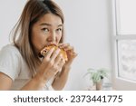 Small photo of Asian Thai woman absent minded, inattentive white having breakfast as homemade sandwich, looking at something or watching tv, thinking about work at home alone on chilling day in the morning.