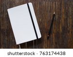 White cover notebook with black pencil on rustic wooden table flat lay photo. Closed notebook with blank cover flat lay photo. Notepad on table top view. Sketchbook banner template. Art logo mockup