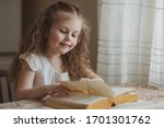 Cute little girl is reading a yellow book at a table by the window at home. The idea of a child's activity in quarantine
