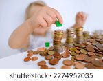 Small photo of small child, blonde girl 3 years old playing with cash, house model on stacks euro currency coins, pocket money, concept home mortgage, home insurance, financial literacy of children, personal savings