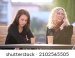 Small photo of Female friends spending time at street cafe outdoors while one girl is talking happily on mobile phone and another waiting frustrated at offensive behaviour of her mate. Friendship problems concept