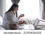 A young woman in a bathrobe works at a laptop in bed.