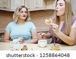Small photo of Young pretty long-haired girl with pleasure drinks tea and eats pancakes with honey. Fat blonde with undisguised envy looks at what is happening. The concept of a healthy diet, diet, weight loss.