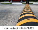 Yellow and Black Striped Speed Bump in Parking Lot with Angled Parking Spaces and Red Car Driving in the Background