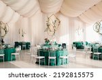 Small photo of The decor of the wedding banquet hall with tables in emerald green, white drapery on the ceiling, gold geometric decorative elements.