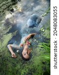 Small photo of Art beautiful romantic woman lies in swamp in blue long dress with flowers. Portrait brunette in transparent dress in water swamp mud duckweed. Book cover