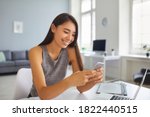 Small photo of Smiling young Asian woman using cell phone in office. Happy millennial girl scrolling through positive comments on social media news feed, chatting online, writing posts for personal blog on mobile