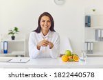 Small photo of Positive dietician sitting at office desk with fruit and glass of water looking at web camera and smiling during client's visit, individual online consultation or while recording video presentation