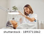 Small photo of Professional female beautician makes a rejuvenating injection to a young woman who lies relaxed with her eyes closed in a modern beauty salon. Concept of smoothing wrinkles with injections.