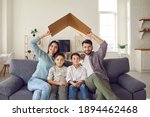 Happy beautiful family of four sitting on couch at home together. Smiling mommy and daddy holding symbolic roof above little kids on sofa. Mortgage, house insurance, future plans, protecting children