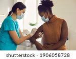 Small photo of Young African American woman getting flu shot during seasonal vaccination campaign. Doctor or nurse in medical face mask cleans skin on patient's arm before injecting modern Covid 19 antiviral vaccine