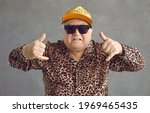 Small photo of Cool retired gangsta granddad flexing to rap music. Portrait of funny weird active rich senior man in baseball cap, trendy eyeglasses and leopard patterned party shirt posing and having fun in studio