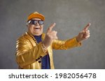 Small photo of Funny happy rich senior man in golden party jacket, baseball cap and bling gold chain necklace enjoying rap music. Old but youthful grandpa dancing and having fun isolated on gray studio background