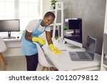 Small photo of Smiley charwoman, caretaker, daily janitor service lady with apron, antiseptic spray bottle and wet cloth cleaning desktop PC computer, laptop, desk and table surfaces in modern office room interior