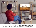 Adult woman takes break from working on laptop and watches movie on TV. Happy black lady resting on sofa in living room interior at home and enjoying favorite show on television set with big screen