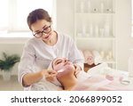 Small photo of Beautician at beauty parlor applying soothing pampering facial mask with pink kaolin clay on young lady's face. Calm beautiful woman relaxing on soft towel while enjoying spa day in modern spa salon
