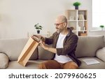 Small photo of Satisfied adult male customer who received package from online store unpacks his order. Man sitting on sofa near consignment note and pulls out cardboard box delivered to him by delivery service.