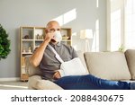 Small photo of Happy bald man with broken arm drinking tea at home. Cheerful male patient who's wearing immobilizer sling after car crash is relaxing on sofa, watching TV, holding mug and enjoying cup of good coffee