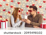 Young man and woman having a fun date on Saint Valentine's Day, sitting at table in cafe or at home, sipping drink from one cup through heart-shaped straws and enjoying cute and funny couple moment