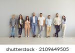 Small photo of Ready to make success happen. Team of happy successful and modern business people standing leaning against gray wall in empty office. Variety of men and women stand in row and look at camera.