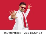 Small photo of Happy smiling good looking brunet man in elegant white suit and party glasses dancing on red background. Studio shot of show presenter, TV commercial actor, singer, dancer or wedding event toastmaster