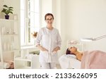 Small photo of Portrait of friendly female woman beautician, aesthetic nurse or masseuse at her workplace. Smiling female beauty salon worker standing next to her client lying on couch in bright office.
