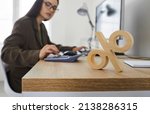 Small photo of Corporate tax and interest rate. Close up of wooden percent sign standing on table on background of serious busy woman counting on calculator. Concept of interest calculation.
