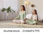 Small photo of Family yoga. Beautiful young woman and her charming little daughter are smiling while doing yoga together at home. Family sits in lotus position on floor in living room. Mom teaches child to meditate.