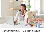 Small photo of Smiling female designer or seamstress talking on webcam giving online consultation to client. Dressmaker sits at her workplace in front of laptop and advises online or conducts sewing master class.