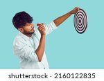 Indian man shooting at target. Profile view of young South Asian man in shirt standing isolated on blue background, holding dart board and aiming little red arrow at bullseye. Setting goal concept