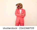 Small photo of Studio shot of strange guy wearing wacky ugly dinosaur mask. Man in pink party suit, bow tie and funny silly scary masquerade lizard mask standing with his arms folded isolated on white background