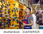 Salesman writing on a clipboard while checking construction and carpentry tools on display. Young man who works at a DIY store or hypermarket looking at various tools on shelves in one of departments
