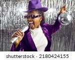 Small photo of Happy young black girl having fun at karaoke party night. Cool excited funky African American woman singer in purple hat, jacket and party glasses holding silver disco ball and singing in microphone