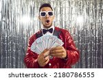 Small photo of Happy wealthy ethnic winner man in shiny suit, bowtie and cool glasses looks at paper money bills bunch with surprised wow face expression. TV game show host guy in foil fringe studio shows prize cash