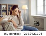Small photo of Tired woman having intense headache. Stressed young Caucasian girl suffering from acute migraine or temporal arteritis pain is sitting on the sofa at home and touching her head with her eyes closed