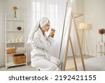 Beautiful young woman in a white dressing gown and bath towel turban on her hair sitting in front of a big mirror in the bedroom, looking at her reflection and applying facial beauty mask on her