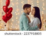 Happy loving couple celebrating St Valentine's Day. Young man and woman enjoying photosession as gift on Saint Valentine's Day, hugging, kissing, dancing in room with balloons and lights in background